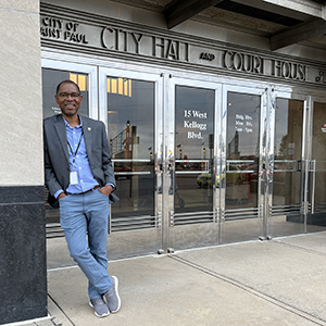 Jules Atangana leans against an outside wall in front of the silver metal Art Deco doors of St. Paul City Hall