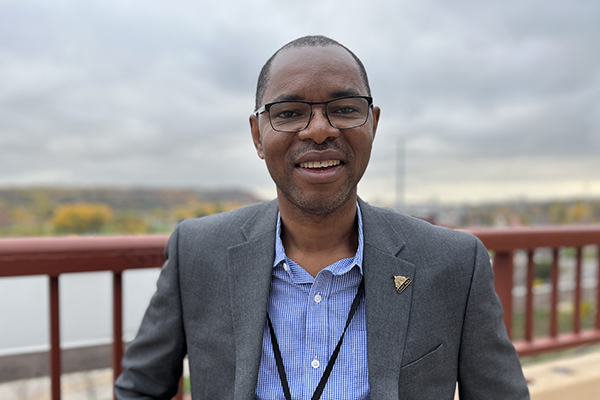 Jules Atangana stands in front of a red metal railing with the Mississippi River its banks of orange and yellow foliage in the background