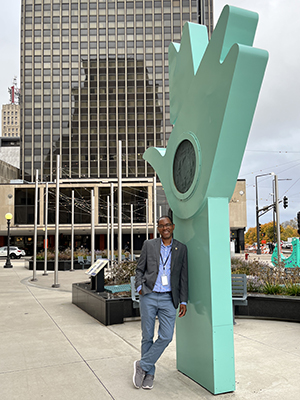 Jules Atangana leans against an outdoor sculpture that resembles a pale green outstretched hand