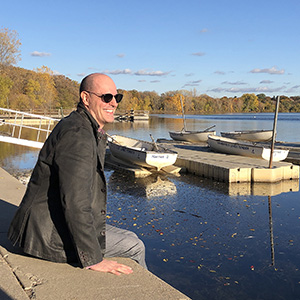 Bald white man wearing a blazer and jeans sits on a concrete ledge overlooking Lake Harriet with boats in background