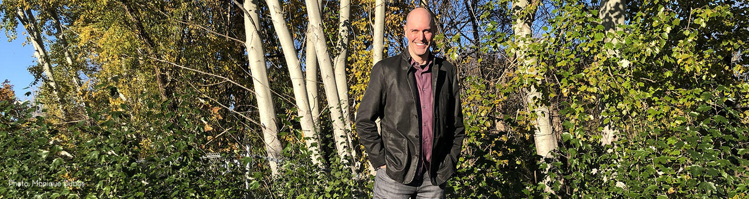A bald white man dressed in blazer and jeans stands in front of a stand of poplar trees in the fall