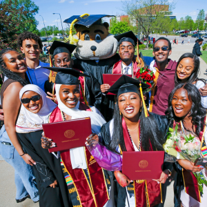 An exuberant group of graduates stand outside in the sunshine with Goldy Gopher following their Commencement ceremony